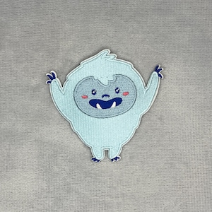以下が含まれることがあります： 腕を上げた水色のイエティのキャラクターが描かれた刺繍ワッペン。イエティは濃い青色の顔、2本の白い歯が見える大きな口、濃い青色の目、小さなピンク色の頬をしています。デザインは白い糸で縁取りされています。