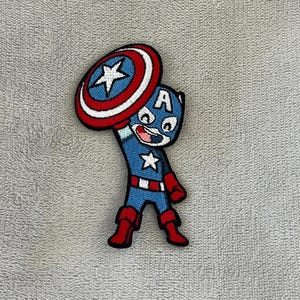 Peut inclure: Écusson brodé représentant le personnage de dessin animé Captain America. Le patch montre Captain America dans un costume bleu et rouge, tenant son bouclier. Le bouclier est bleu, blanc et rouge avec une étoile blanche. Le personnage a un visage souriant.