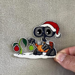 May include: Embroidered patch featuring a cartoon robot wearing a Santa hat, holding a green ornament, and tending a plant with a red ornament. The robot has large, expressive eyes and an orange body. The patch is on a white background.