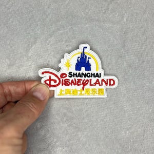 Puede incluir: Parche bordado con el logotipo de Shanghai Disneyland. El parche tiene un fondo blanco con texto y gráficos rojos, amarillos, azules y negros. El parche tiene la forma del logotipo de Disneyland.