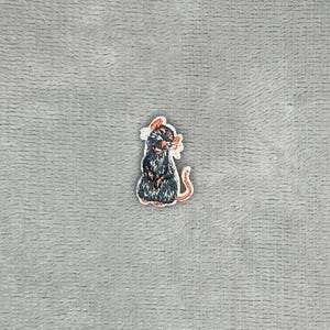 以下が含まれることがあります： 後ろ足で立っている小さなダークグレーのネズミが描かれた刺繍ワッペン。ネズミの耳、鼻、長く曲がった尻尾には淡いピンクのアクセントが施されています。デザインは白い糸で縁取りされており、図形の輪郭がはっきりと強調されています。