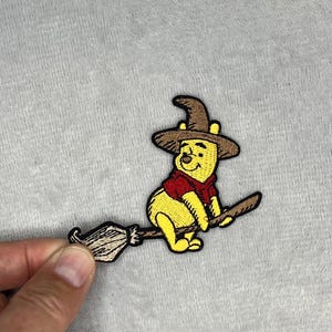 May include: Embroidered patch featuring a yellow bear wearing a brown witch hat and red shirt, riding a broom. The patch has black outlines and is set against a gray background. The bear is smiling.