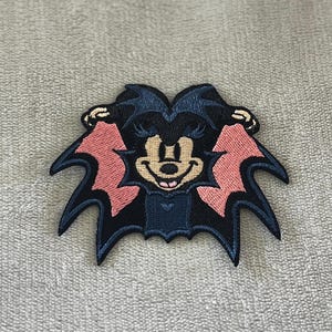 May include: Embroidered patch featuring a cartoon character with bat wings and a bat-like appearance. The patch has a black and pink color scheme with a smiling face. The patch is likely for clothing or accessories.