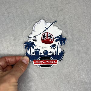 May include: Embroidered patch with a red and white gondola, palm trees, and the word "Skyliner" in a red and white banner. The patch has a blue and white colour scheme and is shaped like a cloud.
