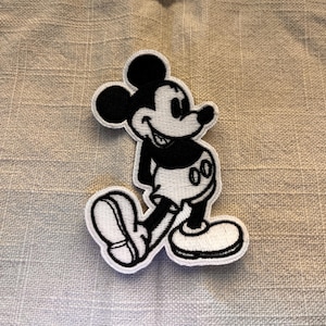 May include: Black and white embroidered iron-on patch of Mickey Mouse. Mickey is smiling and wearing his signature red shorts and yellow shoes.