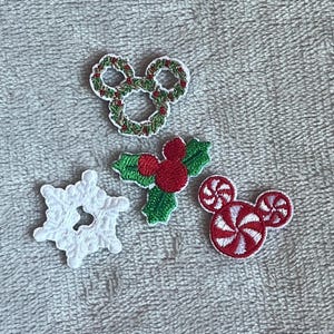 Op de afbeelding: Vier geborduurde patches met vakantiethema's: een groene krans in de vorm van muizenoortjes, een trosje rode hulstbessen met groene bladeren, een witte sneeuwvlok en een rood-wit pepermuntje in de vorm van muizenoortjes.