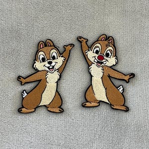May include: Two embroidered patches of cartoon chipmunks. The chipmunks have brown fur, tan bellies, and black outlines. One chipmunk has a red nose. They are in a playful pose with their arms raised. The patches are on a grey background.