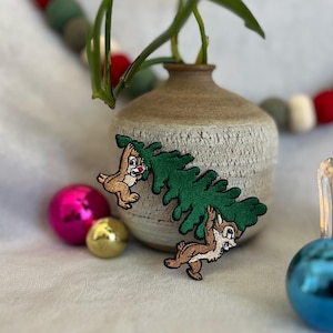May include: Two embroidered iron-on patches featuring Chip and Dale from Disney's Chip 'n Dale Rescue Rangers. The patches show the characters holding a green Christmas tree.