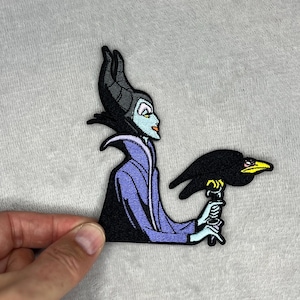 May include: Embroidered patch featuring a stylized depiction of a character with black horns, a purple cloak, and a green face, holding a black raven with a yellow beak. The patch is black, purple, green, and white.