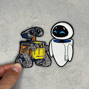 May include: An embroidered patch featuring the characters WALL-E and EVE. WALL-E is a yellow and grey robot with treads, while EVE is a smooth white robot with a black face and blue eyes. The text WALL-E is visible on the yellow robot's chest.