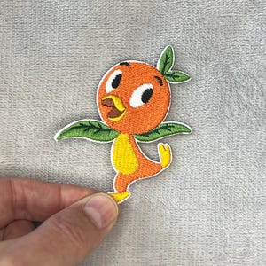 May include: Embroidered patch featuring a cheerful orange bird character with green leaf wings and a yellow belly. The patch has a white border and is designed to be ironed or sewn onto fabric. The bird is in a playful pose.