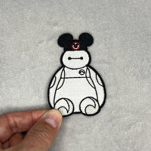 May include: Embroidered patch of a white, round character with black outlines and details. The character has a black head with Mickey Mouse ears and a red circle emblem. The patch is held against a grey background.