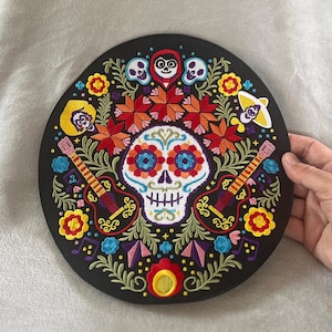 Può includere: Una toppa circolare nera ricamata con un designo di teschio di zucchero colorato, circondato da fiori, foglie e note musicali. Il teschio ha un viso bianco con occhi neri, un naso rosso e una bocca rosa. Il teschio è circondato da un design floreale colorato con fiori rossi, arancioni, gialli, verdi, blu e viola. La toppa è perfetta per aggiungere un tocco di cultura messicana a qualsiasi progetto.