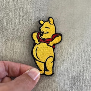 May include: Embroidered patch featuring a yellow bear with a red scarf. The bear is smiling and has its arms outstretched. The patch has a black border and is likely for clothing or accessories.