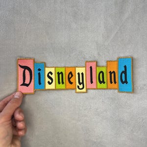 Könnte beinhalten: Bunter Disneyland-Aufnäher mit dem Wort "Disneyland" in schwarzer Schrift. Die Buchstaben sind vor einem Hintergrund aus rosa, blauen, gelben, grünen und orangefarbenen Rechtecken mit einem goldenen Rand angeordnet. Der Aufnäher wird von einer Hand gehalten.