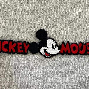 May include: Embroidered patch featuring Mickey Mouse. The patch has the words "MICKEY" and "MOUSE" in red, with a black outline. The iconic character's face is in the centre, with a white face, black ears, and a red smile.