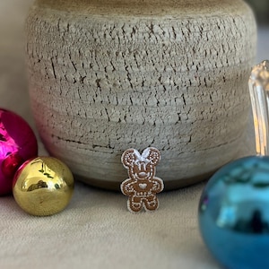 Puede incluir: Una galleta de jengibre marrón con forma de oso con detalles de glaseado blanco. La galleta está sobre una superficie blanca con un jarrón y adornos navideños en el fondo.