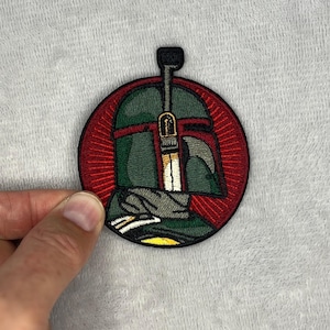 Pode incluir: Patch bordado com um design de capacete verde e cinza dentro de um círculo vermelho. O patch tem uma borda preta e uma representação detalhada das características do capacete. O design lembra um personagem popular de ficção científica.