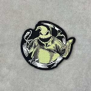 Op de afbeelding: Een zwart-witte patch met een cartoonfiguur met een groen gezicht en lichaam. Het figuur heeft een gestikte textuur. De patch is cirkelvormig met witte accenten.
