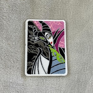 May include: Embroidered patch featuring a stylized illustration of a character with black horns, a green tongue, and a black and white outfit. The background is pink with a floral pattern. The patch has a white border.