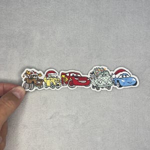 May include: An embroidered patch featuring a row of five cartoon vehicles with holiday themes, including a brown truck with antlers, a yellow car, a red race car, a green van carrying a Christmas tree, and a blue car, all wearing Santa hats.