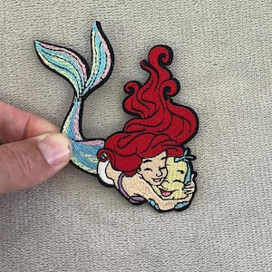 Op de afbeelding: Geborduurde patch met een zeemeermin met rood haar en een kleurrijke staart, die een lachend personage omhelst. De patch heeft een zwarte rand en is geschikt voor kleding of accessoires.