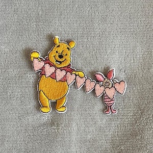 Könnte beinhalten: Bügelbild mit Winnie Puuh und Ferkel, die eine Girlande aus rosa Herzen halten.