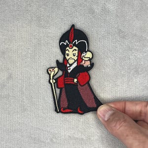 May include: Embroidered patch featuring a cartoon depiction of Jafar from Aladdin. The patch showcases Jafar in his signature black and red attire, holding a staff with a snake head. A small parrot is perched on his shoulder.