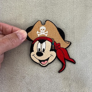 May include: Embroidered patch featuring a cartoon mouse with a pirate hat and red bandana. The tan pirate hat has a skull and crossbones emblem. The mouse has a smiling expression and is wearing a red bandana with beaded accents.