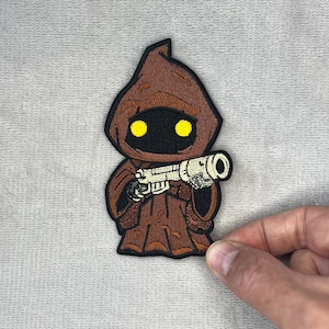 May include: An embroidered patch depicting a cartoon Jawa character from Star Wars. The Jawa is brown and black, with yellow eyes and a hooded robe. It is holding a silver blaster.