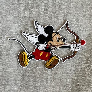 Puede incluir: Un parche bordado para planchar con Mickey Mouse vestido de Cupido, con alas, sosteniendo un arco y una flecha con una punta de corazón roja.
