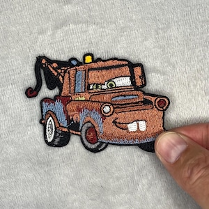 Può includere: Toppa ricamata raffigurante il personaggio di Mater del film Cars. La toppa è principalmente marrone con dettagli blu e rossi, che mostrano il design del carro attrezzi di Mater. La toppa ha una faccia sorridente e pneumatici bianchi.