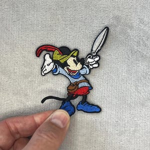 May include: Embroidered patch featuring a cartoon mouse character in a medieval outfit, holding a silver knife. The patch has a black, red, blue, green, and white color scheme. The mouse is wearing a hat and boots. The patch is on a gray background.