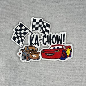 May include: An embroidered patch featuring a brown tow truck and a red race car with a yellow lightning bolt design. Two black and white chequered flags are positioned above the vehicles, and the text KA-CHOW! is embroidered in black letters.