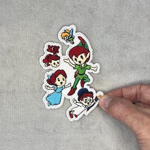 May include: An embroidered patch featuring a group of cartoon characters including a boy in a green outfit, a small fairy with yellow hair, a girl in a blue dress, a girl with red hair, and a character in a white coat holding a yellow umbrella.