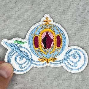 May include: Embroidered patch featuring a light blue and white Cinderella carriage design. The carriage has gold accents, a pink interior, and a dark red door. The patch is held by a hand.