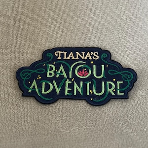 May include: A dark blue embroidered patch with green vines and a red flower. The text on the patch reads "Tiana's Bayou Adventure".