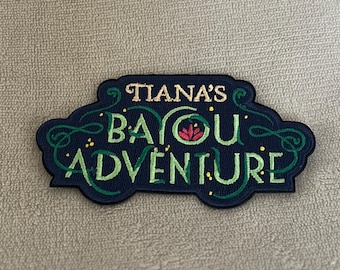 May include: A dark blue embroidered patch with green vines and a red flower. The text on the patch reads "Tiana's Bayou Adventure".