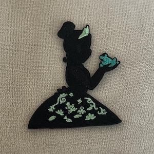 Może przedstawiać: Czarna i zielona haftowana naszywka do prasowania przedstawiająca sylwetkę kobiety trzymającej żabę. Sukienka kobiety jest ozdobiona wzorem kwiatowym.