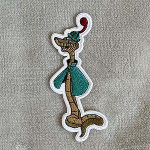 May include: An embroidered patch of a cartoon snake wearing a green cape and a red and green hat. The snake is brown and has a long, thin body.