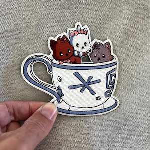 Op de afbeelding: Geborduurde patch met drie cartoonkatten in een theekopje. Het theekopje is wit met een blauwe omtrek en handvat, versierd met een blauwe asterisk en andere kleine ontwerpen. De katten zijn bruin, wit en grijs.