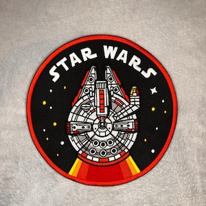 Może przedstawiać: Okrągła naszywka z statkiem kosmicznym Millennium Falcon z Gwiezdnych Wojen. Naszywka ma czarne tło z białym napisem "STAR WARS". Statek kosmiczny jest szczegółowo przedstawiony w kolorach czerwonym, białym i szarym, z żółtymi i białymi gwiazdami.