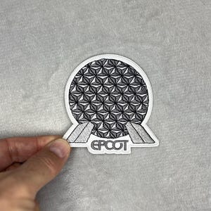 May include: A white and black patch featuring the iconic geodesic sphere design with the word "EPCOT" in a gray font. The patch is held against a gray background.