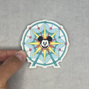 Puede incluir: Un parche bordado circular con un diseño de noria estilizada en azul claro, turquesa y blanco. En el centro, un patrón de estrella amarilla rodea una cara de Mickey Mouse en blanco y negro con expresión sonriente y detalles rojos.