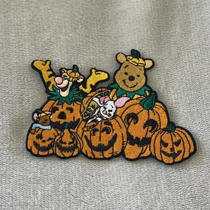 May include: Embroidered patch featuring Winnie the Pooh, Tigger, and other characters atop carved orange pumpkins. The patch has a black border and is set against a neutral background. The patch is a Halloween-themed item.