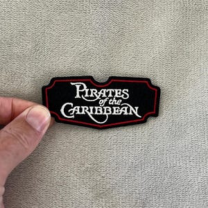 Puede incluir: Parche bordado negro con las palabras "Pirates of the Caribbean" en escritura blanca. El parche tiene un borde rojo y se muestra sobre un fondo neutro.