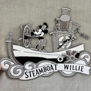 May include: Embroidered patch featuring Mickey Mouse at the helm of a steamboat. The black, white, and gray design includes the text "Steamboat Willie" on a banner. The patch is likely for clothing or accessories.