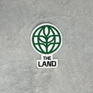 Peut inclure: Écusson brodé blanc et vert représentant un globe avec un motif de feuilles. En dessous, le texte "THE LAND" en noir sur un rectangle blanc. L'écusson est posé sur un fond gris texturé.