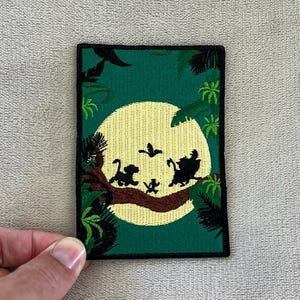 Puede incluir: Parche bordado con una escena de la jungla con una gran luna amarilla y siluetas de personajes. El parche tiene un fondo verde con bordes y acentos negros, y es de forma rectangular.