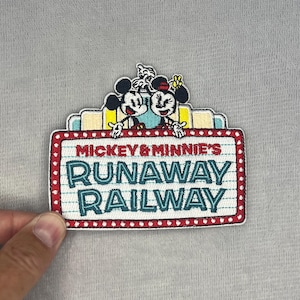 May include: An embroidered patch featuring Mickey and Minnie Mouse above a marquee sign that reads MICKEY & MINNIE'S RUNAWAY RAILWAY in red and teal lettering. The sign has a white background with thin blue lines and a red border with white dots.